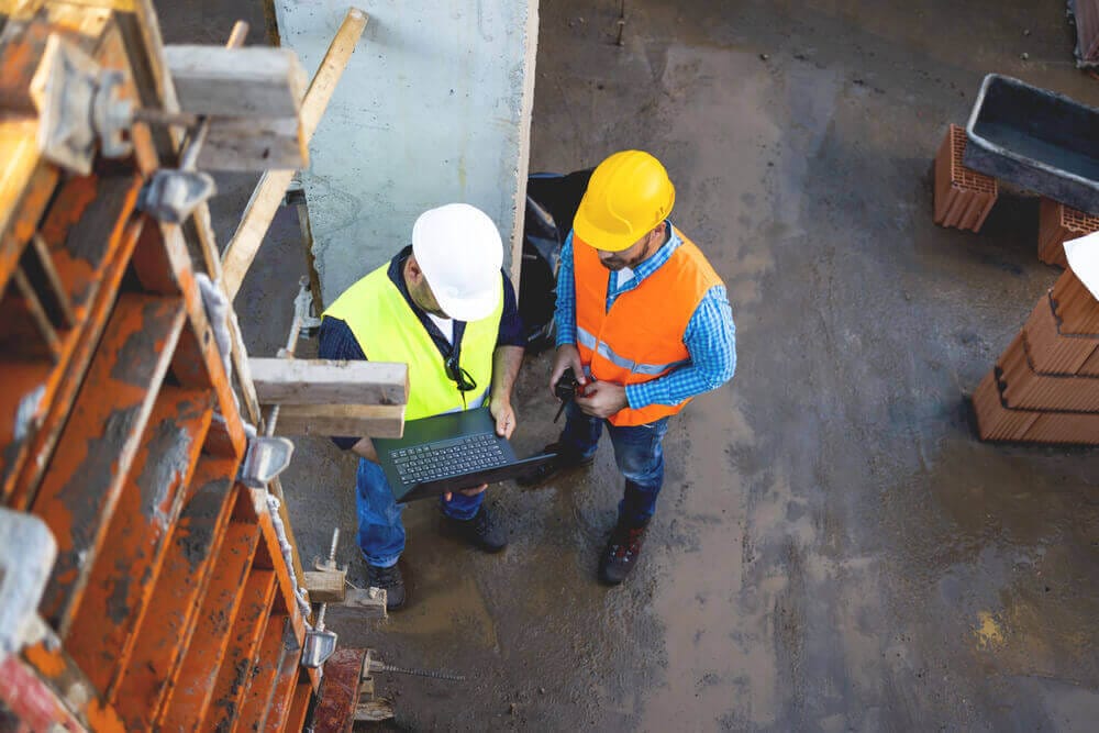 Dois trabalhadores em um canteiro de obras, um ao lado do outro, ambos equipados com capacetes e coletes de segurança.